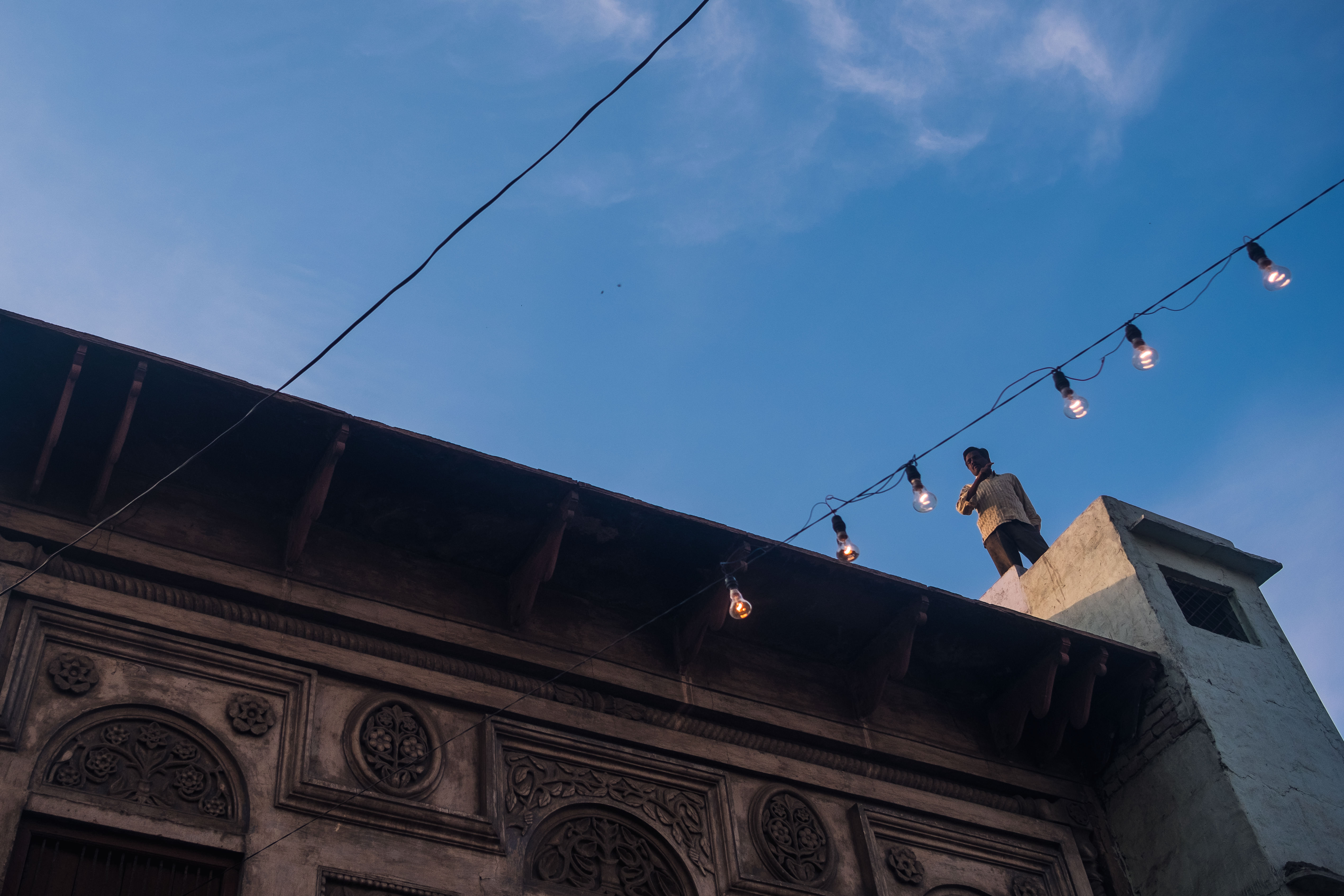 India Street Photography During Holi Festival. man stand on the roof between string lights. Images by Jason Vinson